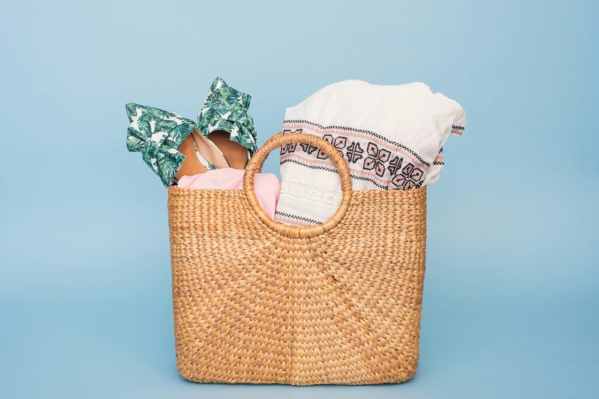 A woven straw tote bag filled with beach essentials, including a tropical-print swimsuit and embroidered cover-up, set against a light blue background.

